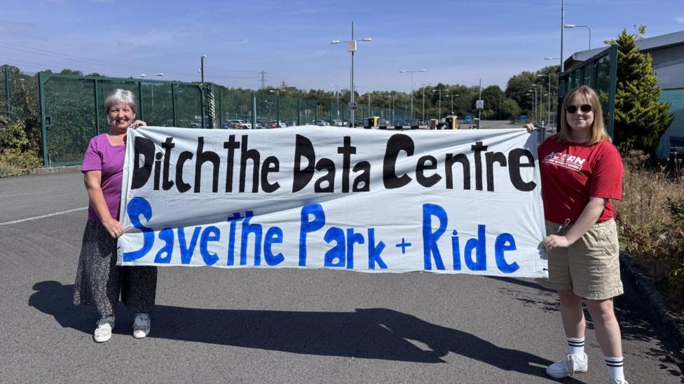 Two campaigners smiling holding a homemade banner which says "ditch the data centre, save the park & ride"