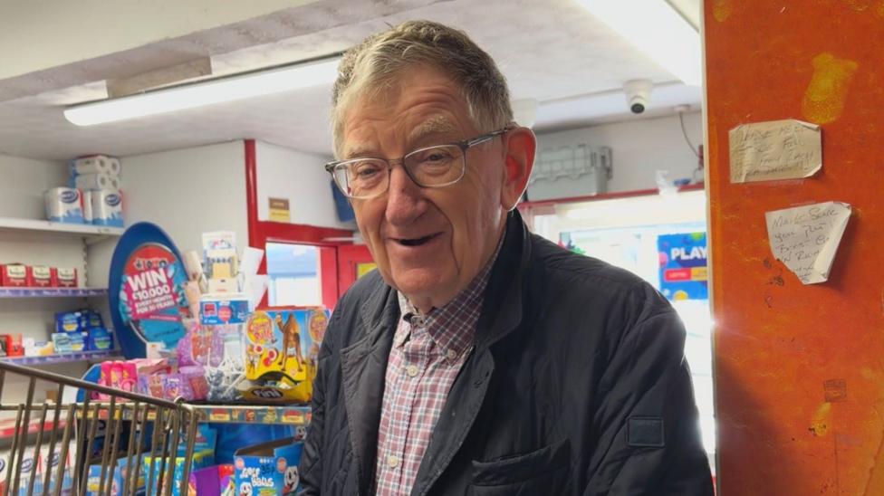 Lyle Cubitt stands in Diamonds, he has grey hair and glasses and is wearing a dark blue jacket over a red, white and blue checked shirt.