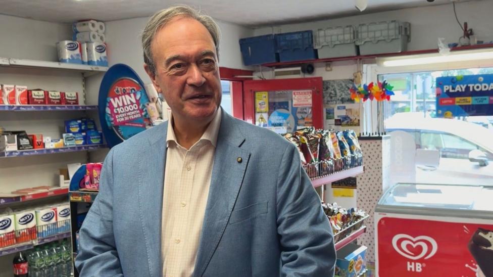 Carroll Falls stands in Diamonds newsagents. He has grey hair and is wearing a light blue suit jacket over a white shirt.