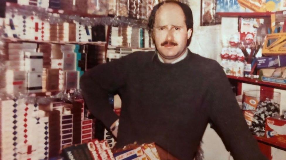 Eugene Diamond in his shop in the early 1980s. He has brown hair and a brown moustache and is wearing a dark green jumper. Behind him are stacked packets of cigarettes.