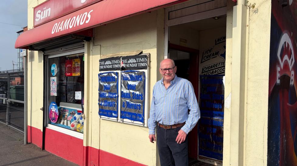 Eugene Diamond stands outside his shop. He is bald and is wearing black-rimmed glasses and a pale blue shirt with yellow stripes. The shop is painted cream with a red section along the lower part of the wall and a red sign reading "Diamonds" in white font.