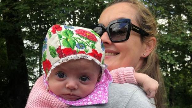 Baby Mairi being carried Maeve, wearing a colourful bonnet and looking over her mum's shoulder at the camera