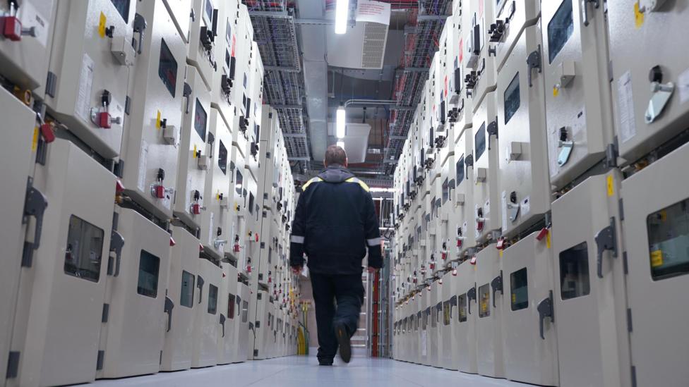 A man in dark jacket walks between two rows of beige coloured metal boxes under strip lighting in the high voltage switching room of a London data centre
