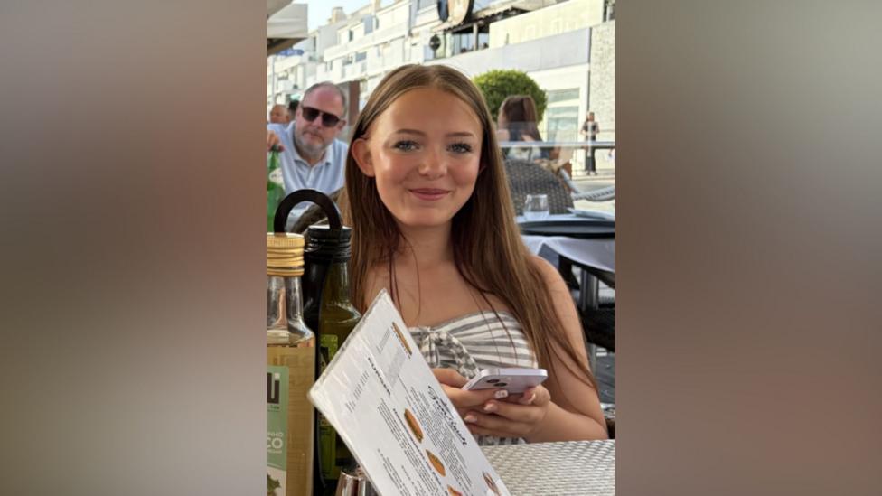 Lillie Nichols has a horizontal striped top and is sitting at a restaurant with a bottle of olive oil on the table, a menu and is holding a white smart phone.