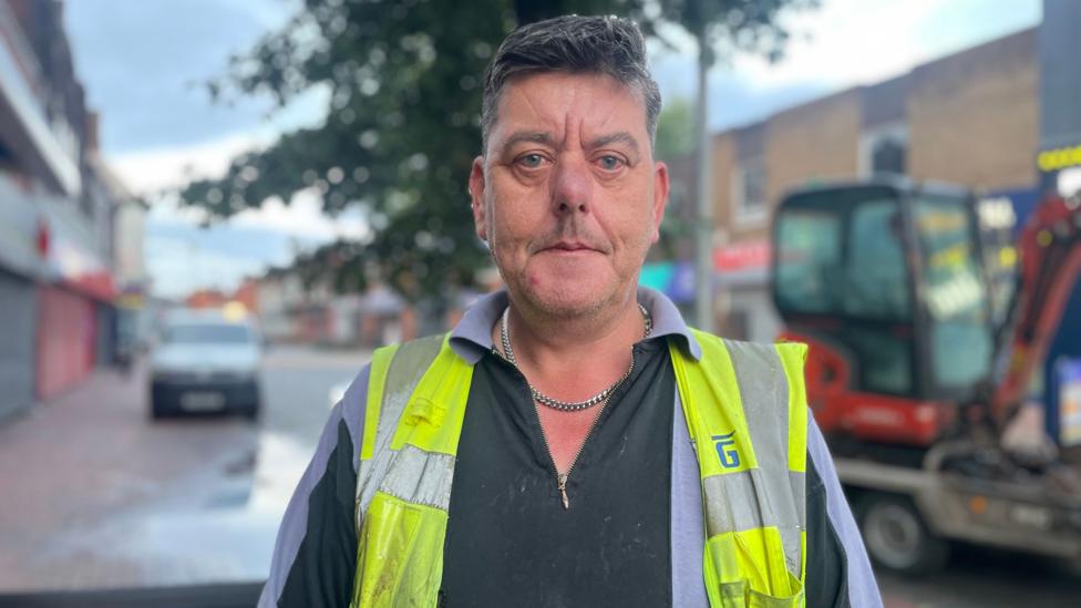 A man wearing a black and grey long-sleeved top with a yellow hi vis over and a silver chain, with dark hair, stands in a street with a red digger and a van behind him and water on the floor.