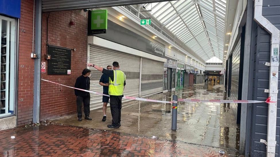 An arcade with shops shuttered up along either side, and three people, including a man in a hi-vis vest, stand by tape closing the walkway off.