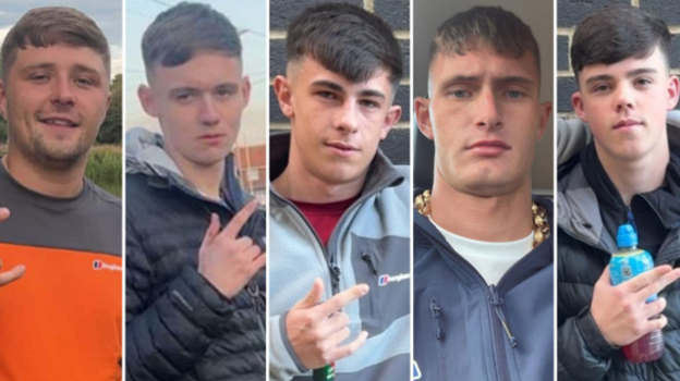 Five young men in their late teens and early 20s. They all have similar haircuts, with a fringe and faded back and sides. Four of them are gesturing with their fingers. Three of them extend their thumb, index, and pinky fingers - leaving their middle and ring finger tucked down. One points with his index and middle finger whilst holding a Lucozade bottle. They are all dressed in casual clothing.