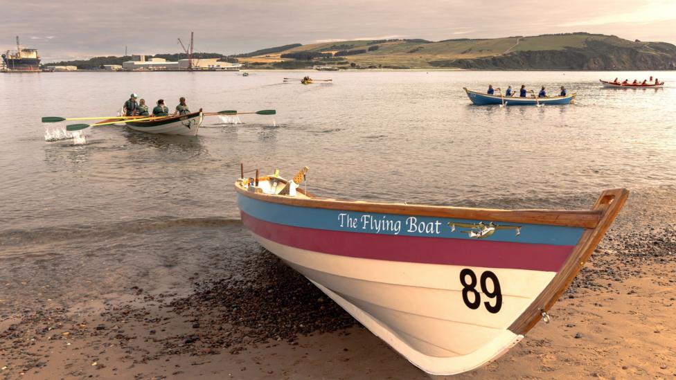 A painted skiff named The Flying Boat rests on a sandy shoreline while several crews row traditional skiffs across calm water during a regatta.
