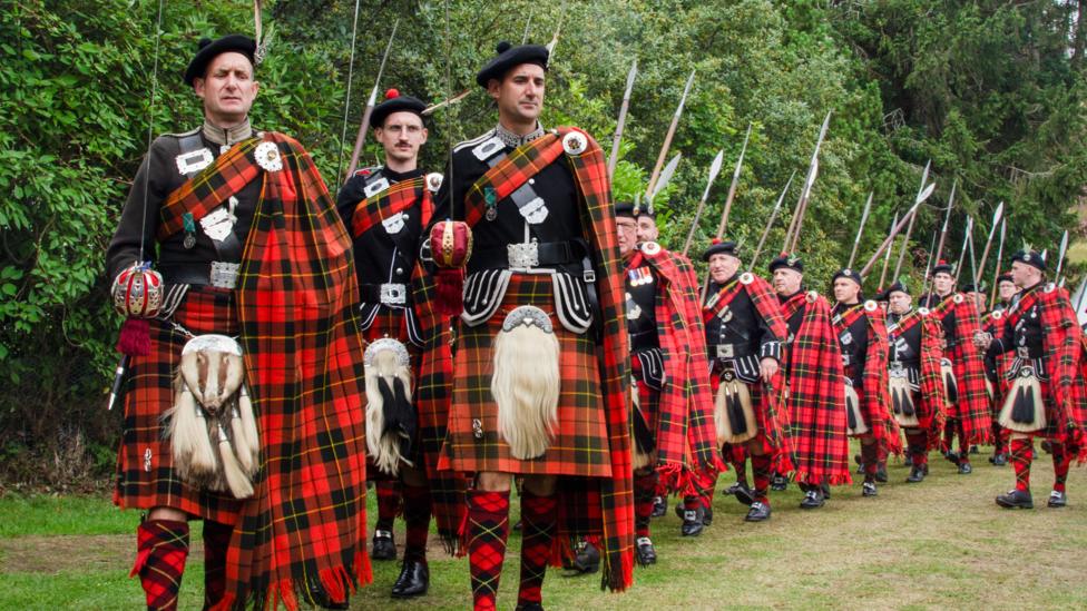 A procession of Lonach Highlanders in traditional tartan dress marches across a grassy field, carrying long pikes and wearing matching red and black tartan plaids.