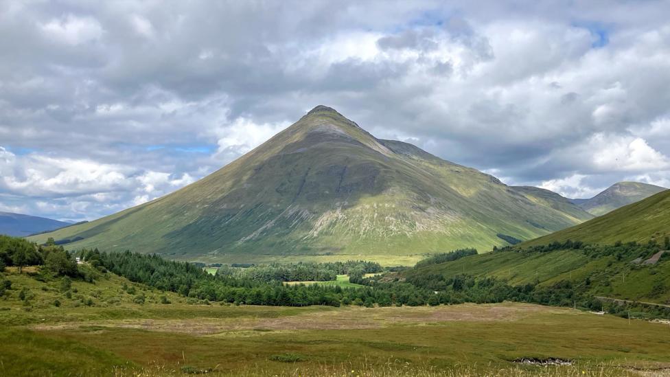 A dramatic conical mountain rises above a green valley, illuminated by patches of sunlight beneath a sky filled with large grey and white clouds.