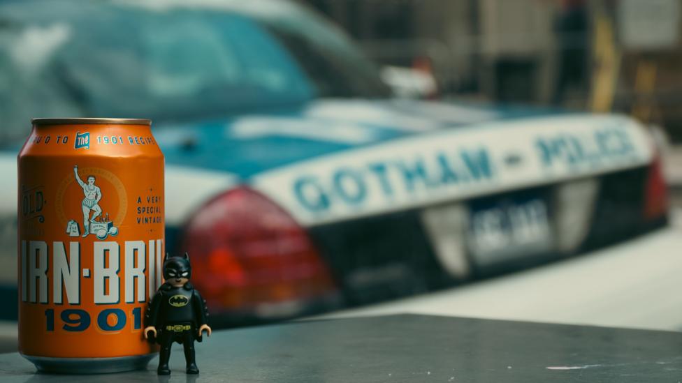 A miniature Batman figure stands beside a vintage can of Irn-Bru in the foreground, with a blurred Gotham City Police car visible in the background.