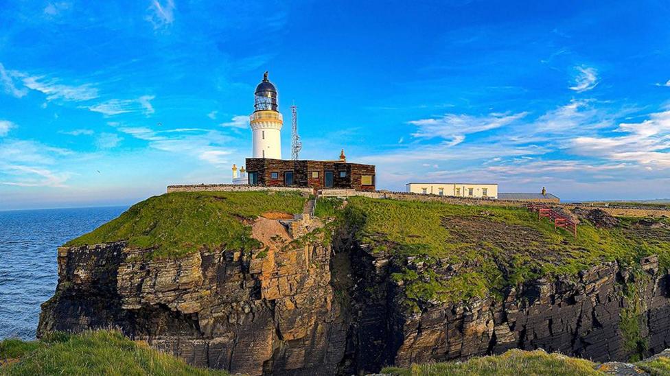 Noss Head Lighthouse stands on a grassy cliff-top headland overlooking the sea.