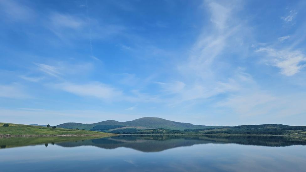 A calm view across Clatteringshaws Loch, where green hills and white clouds are reflected almost perfectly in the still water.