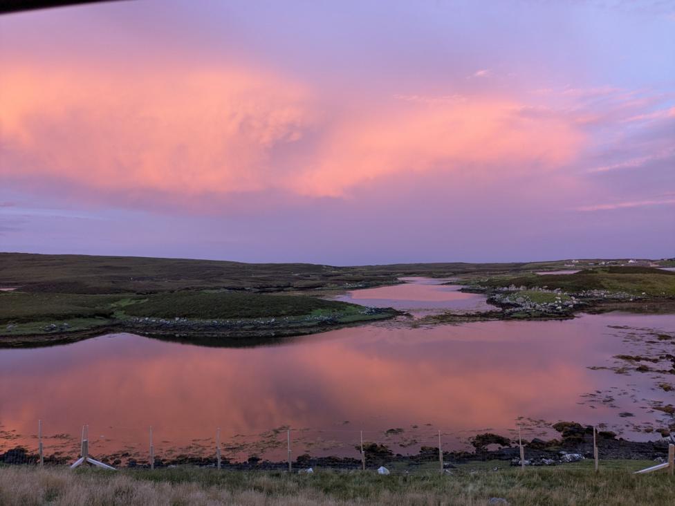 A calm tidal inlet at Locheport on the Isle of North Uist reflects soft pink and purple sunrise colours across the water.
