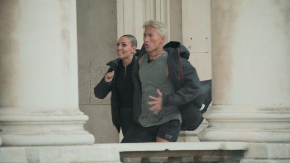 Carlos Gu and Amy Dowden pictured running side by side during an episode of the show, Celebrity Hunted. Carlos is wearing a grey dark coat, a light grey t-shirt and several necklaces. Amy is wearing a black top, jacket and coat. They are both not looking at the camera.