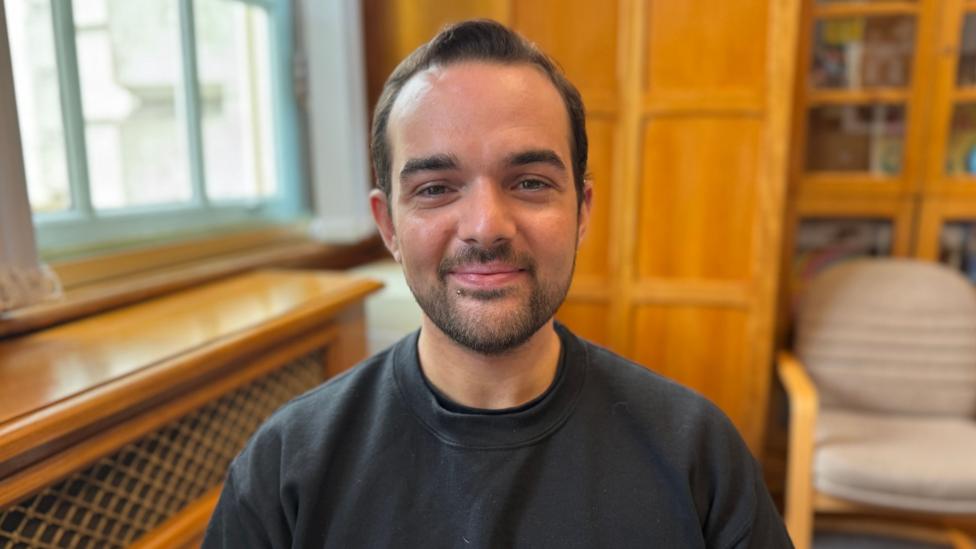 Councillor Micky Murray is a young man with short, black hair and a beard. He is wearing a black jumper and is smiling at the camera. He is sat in an office with wooden bookcases and radiator covers around him.