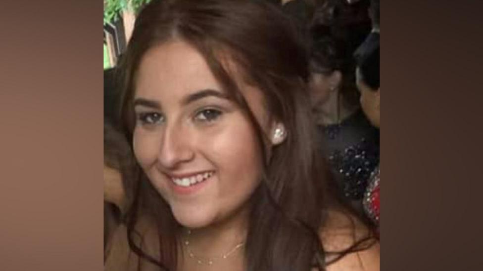 A photo of Georgia Jones from a night out. She is a young woman with long, dark brown hair. She is smiling at the camera. There are blurred lines at either side of the image as it is cropped.