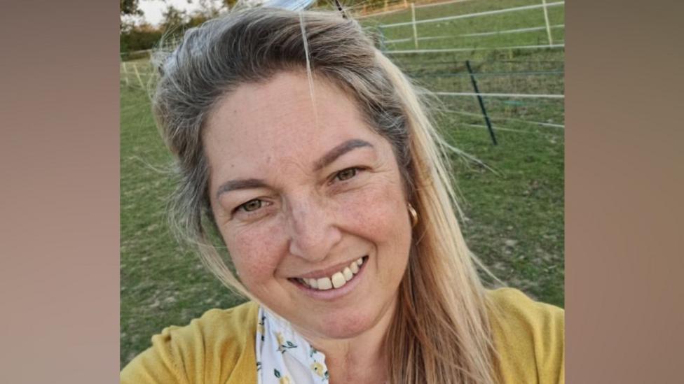 Janine Milburn is a woman with blonde hair. She is smiling with her teeth showing as she takes a selfie. She is standing in a field and is wearing a yellow cardigan and a floral top. 