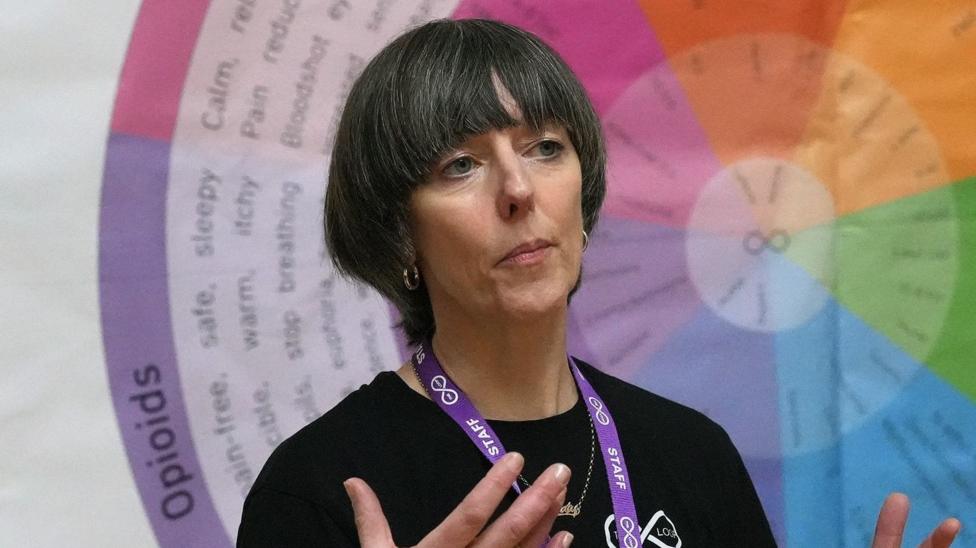 Katy Porter is standing giving a speech in front of a large, colourful wheel in the background. She has short, greying hair and is wearing a purple staff lanyard and a black t-shirt.