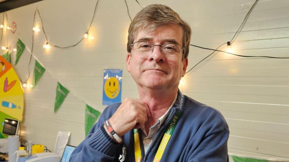 Prof Eamon Keenan is an older man who is wearing rectangular, frameless glasses and is looking at the camera with a neutral expression. Behind him are some string lights, and some green flag bunting. She is wearing a blue jumper and a white shirt.