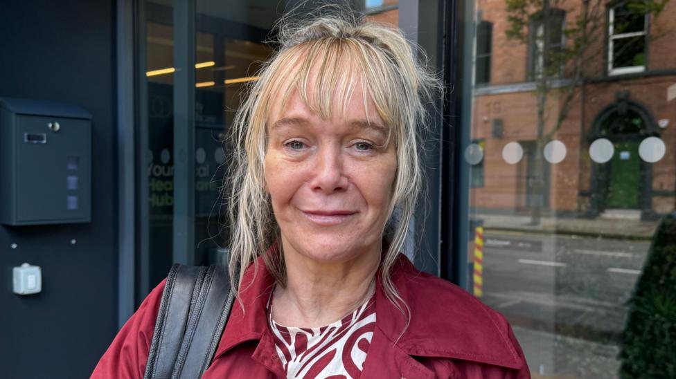 Prof Anne Campbell is blonde with her hair tied up apart from her fringe. She is smiling at the camera. She is wearing a red coat, a red and white patterned shirt and has a black bag strap over her shoulder.
