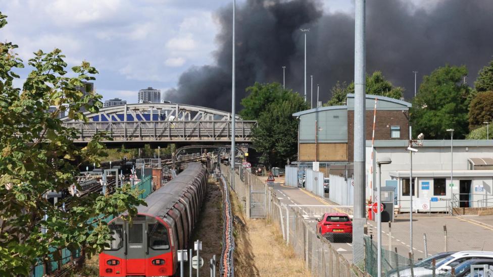 A thick plume of dark grey smoke rises into the sky behind a modern multi-storey building, overhead line gantries, and a arched railway bridge.