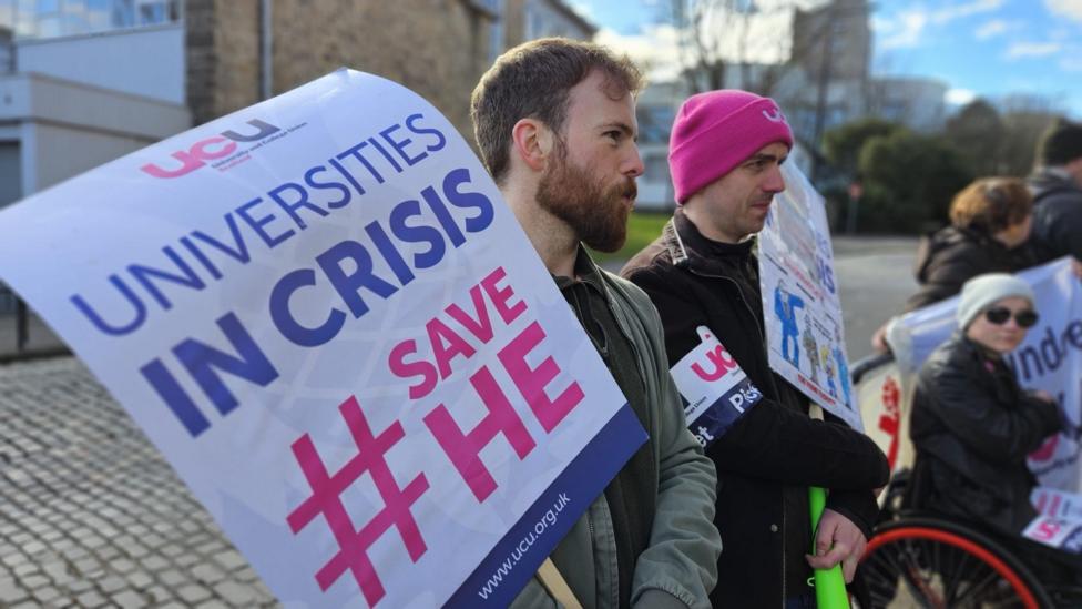 A protester with a beard holds a banner saying Universities in Crisis with two other protesters, one in a wheelchair, in the background