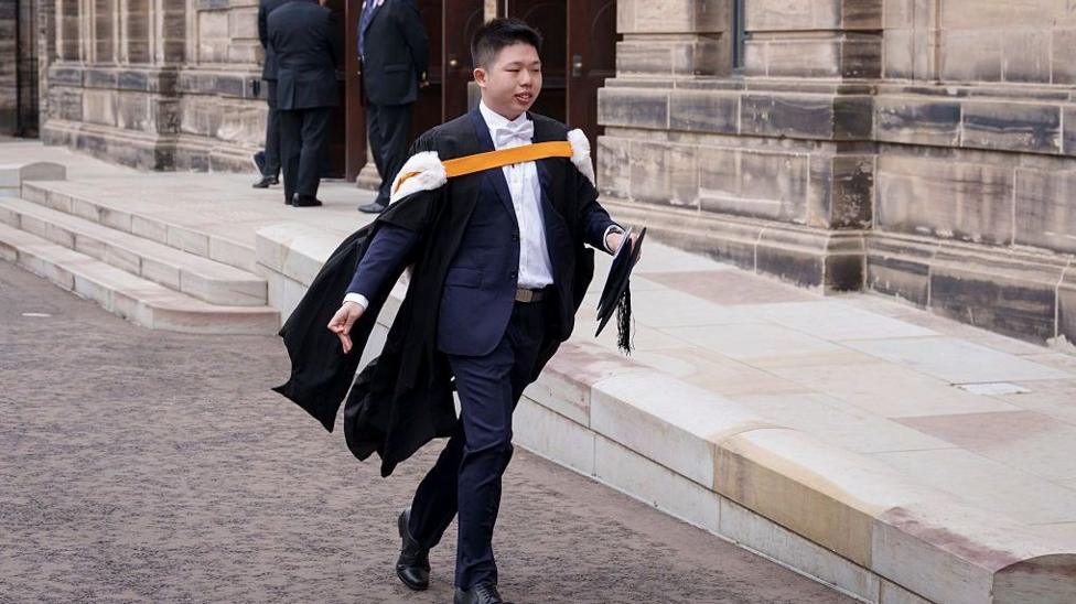 A man in a dark suit and academic gown walks outside a stone university building, carrying a mortarboard cap and documents, with steps and other people visible in the background.