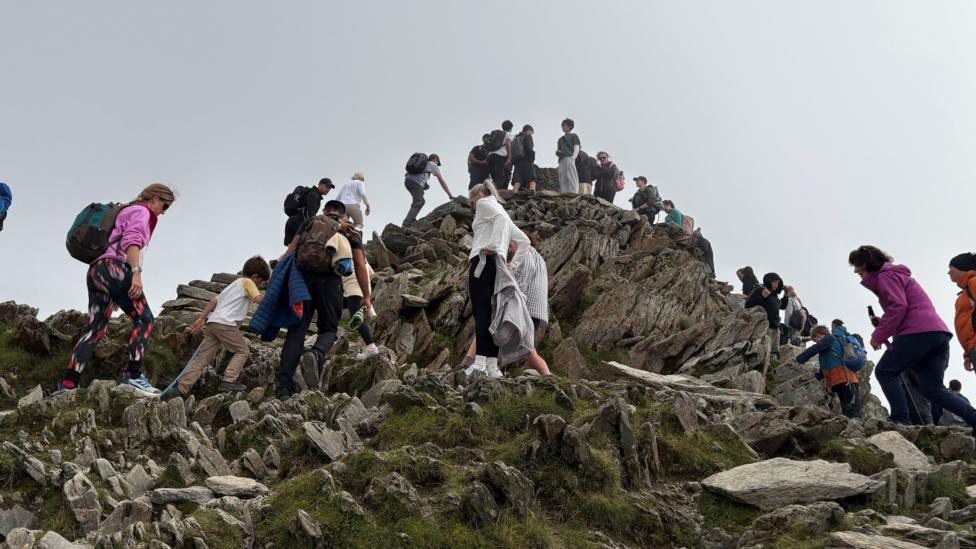 A busy mountain summit is packed with walkers navigating uneven rocky ground. People are spread across the ridge in all directions beneath a blanket of low cloud.