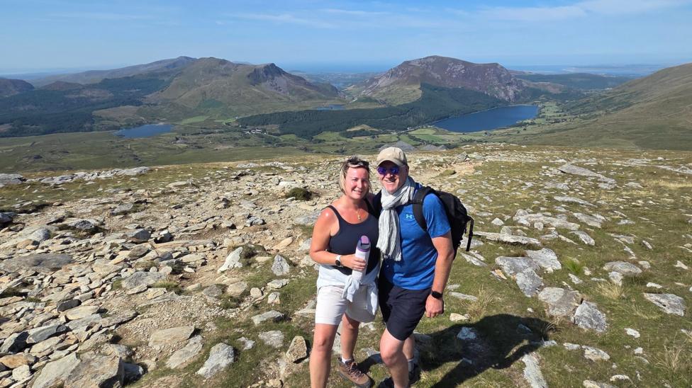 Serena and Patrick smile at the camera. Behind them is an expansive mountainous backdrop. Lakes are visible and forested areas. Serena has blonde hair and is wearing a navy tank top and white shorts. Patrick is wearing a grey cap and blue t-shirt and navy shorts.