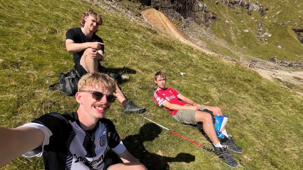 Three young men sit on a grassy hillside during a mountain walk. James Munday takes a selfie in the foreground wearing a black and white sports shirt and sunglasses. He has blonde hair and a moustache. Two others sit higher up the slope. Stan sits highest on the hill and has a blonde mullet and is wearing a black shirt and shorts. Fin lays next to them wearing a red shirt and kaki shorts. He has short brown hair. Walking poles, backpacks and rocky mountain scenery can be seen around them.