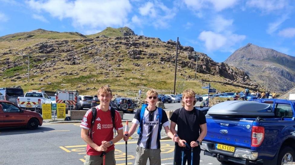 Three walkers stand beside a low stone wall at a mountain car park before starting their last hike. The foreground is filled with stonework and road markings, while rugged hills and a prominent mountain peak dominate the background under bright sunshine.