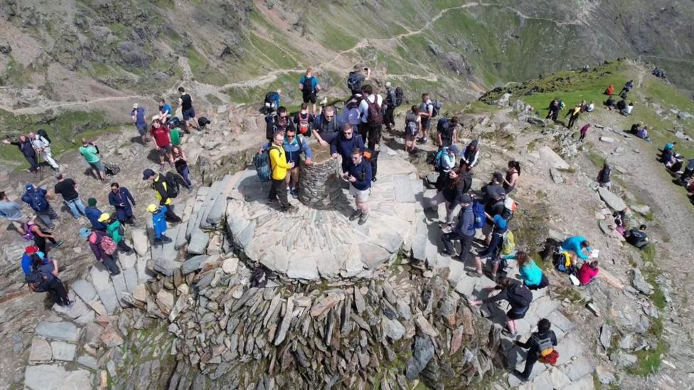 A drone shot of a crowded mountain summit, packed with hikers. People stand, sit and take photographs around a stone summit marker, with winding mountain paths and steep slopes stretching into the distance.