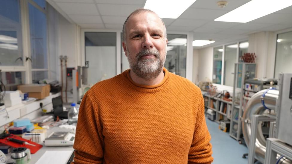 Professor Huw Morgan is stood in a large lab with a lot of equipment behind him. He is looking at the camera with a wry smile on his face. He has a beard and short hair and is wearing a rusty orange sweater.