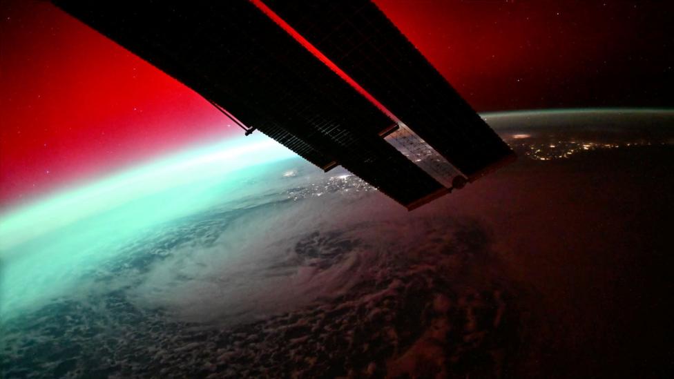 An image of the curvature of the earth taken from space. The sky is a deep red, with part of the earth in light, while most of it is clearly in darkness, with some flashes of lights to be seen. It's not possible to see where it is, as there's a lot of cloud cover. In the foreground is a part of the space station which looks like a wing.