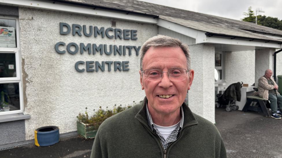 Man with grey hair and glasses. He is standing in front of a white building with grey text on it saying 'Drumcree Community centre'. He is wearing a green quarterzip with a black checkered shirt and white t-shirt beneath.