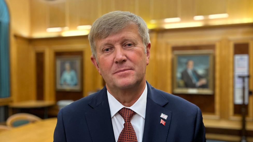 Peter Harris looking at the camera while standing in a wood-panelled council chamber. He is wearing a suit and tie. The tie is maroon and the blazer is navy blue. There are portraits on the wall in the background. He has two metal pin badges on a lapel - one of the union flag and one of the Essex triple seaxes.