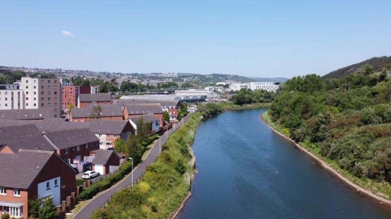 The River Tawe surrounded by green trees with a footpath on the side of a river and houses.