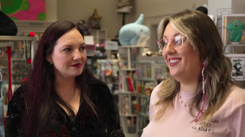 Two women in a shop. One has dark hair and red lipstick and the other has blonde hair and pink glasses.