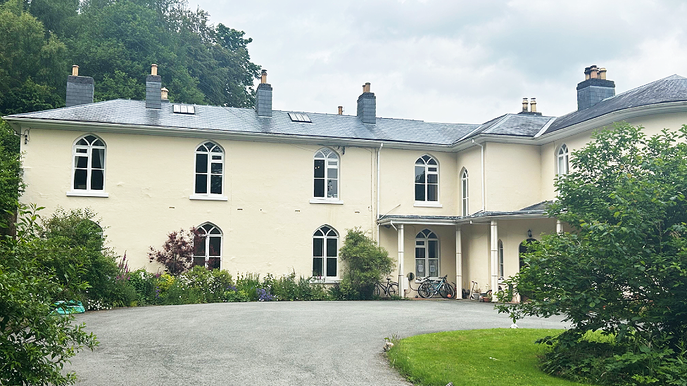 The hall is an early 19th century building. It is two-story light cream colour, with 10 arched upper and lower windows visible along one wall which have curved white-painted leaded patterns across the glass. A covered porch with the entrance door is just visible behind a hedge in front of a curving driveway. There are two skylights in the roof. There are flowering shrubs planted in front of and to the left of the hall. Some bicycles are visible underneath the porch. It looks well cared for.