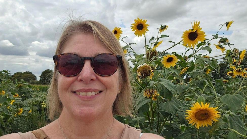 A woman with light brown hair wearing brown sunglasses smiling, she is stood in front of a sunflower field. 