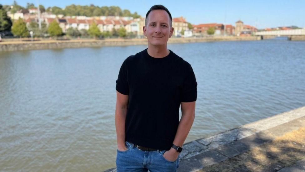 A man with dark short hair wearing a black top and blue jeans smiling infront of a river bank. 