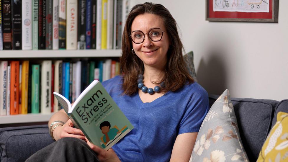 A woman with brown hair and glasses smiling, she is wearing a dark blue top with a blue necklace and is sat on a grey sofa holding a book. 