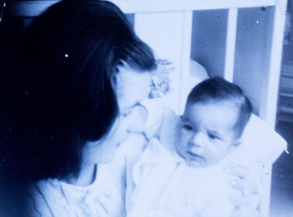 An old, black and white photograph of a young woman cradling a baby. They are crouched next to a cot.