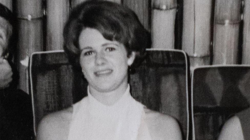 A black and white picture of a young woman in a white, sleeveless top. She is sitting on a chair in front of curtain.