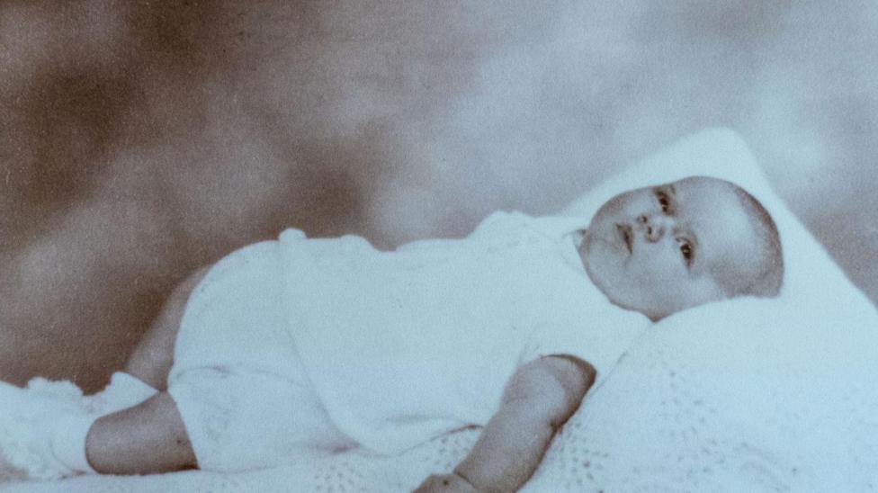 A photograph of a baby dressed in white and lying on white blanket