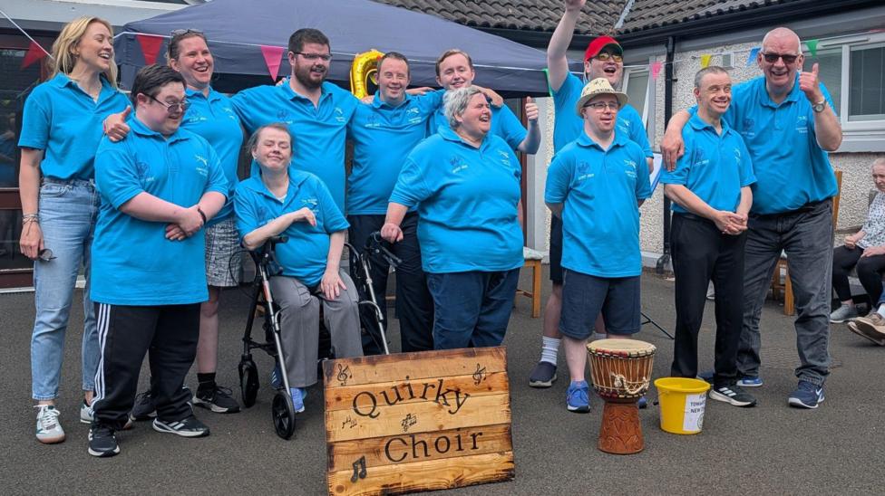A group of people in matching bright blue polo shirts stand together outdoors at a community event, posing behind a wooden sign that reads “Quirky Choir.”