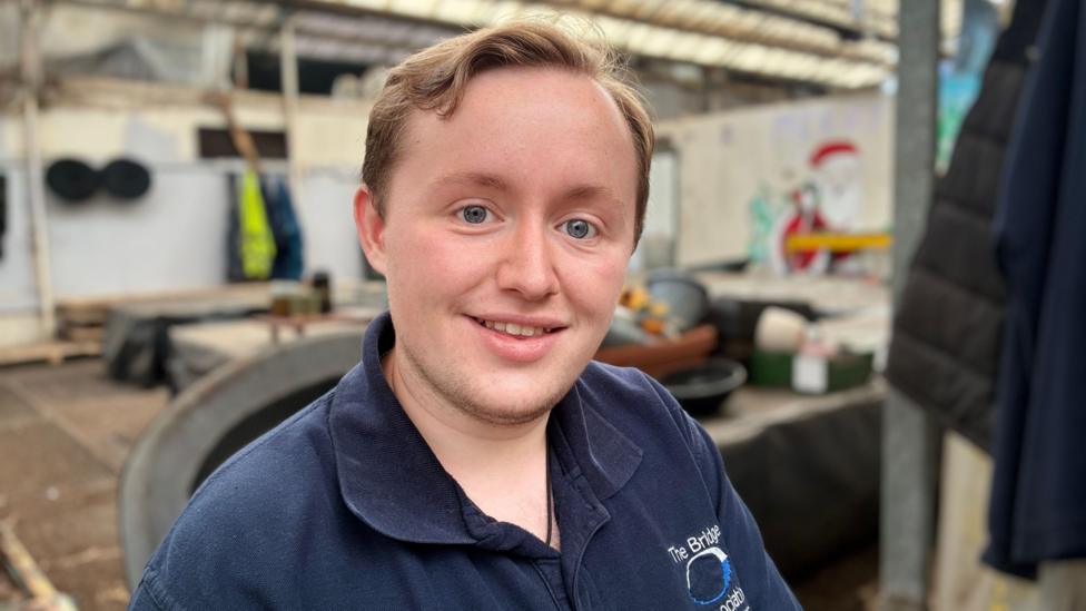 A young man with light brown hair and blue eyes smiles at the camera. He’s wearing a navy-blue collared shirt embroidered with “The Bridge Association” There is a workshop space with tools, containers, and creative wall art in the background. 