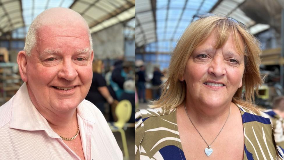 Man and woman smiling at camera. Man is wearing a pink shirt and woman a tropical green top. The man has a chain around his neck and the woman has a heart necklass. There is a workshop area blurred in the background. 