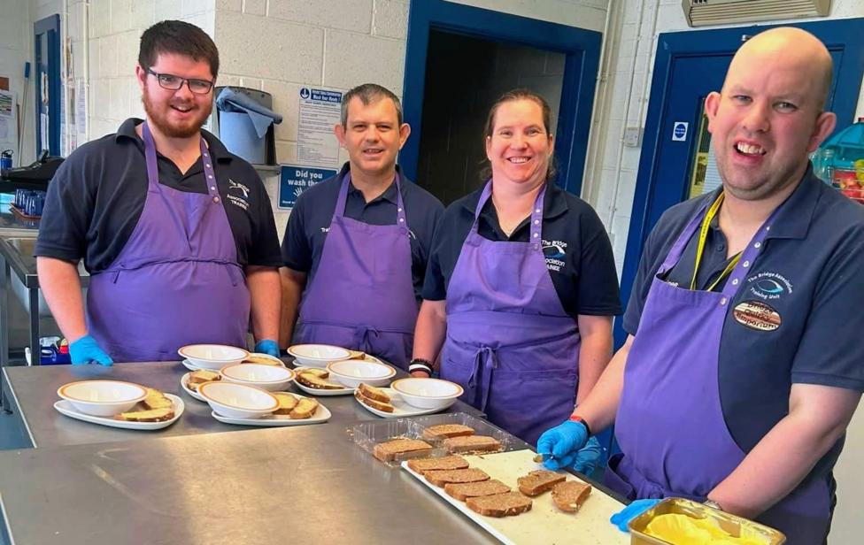 Four people wearing purple aprons and blue gloves prepare sandwiches and meals in a commercial kitchen, standing behind a stainless-steel counter with plates and bowls ready for serving.